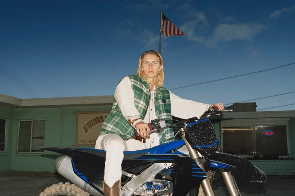 A blonde male fashion model poses on a blue dirt bike in front of Emma’s Holland restaurant with the American flag waving overhead.