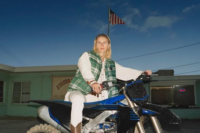 A blonde male fashion model poses on a blue dirt bike in front of Emma’s Holland restaurant with the American flag waving overhead.