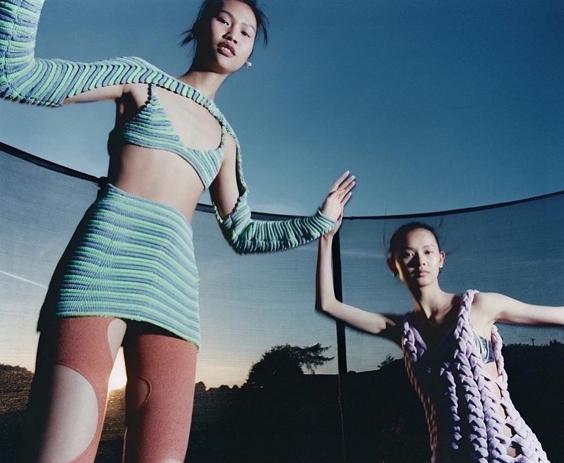 Two women pose outdoors wearing knitwear against a blue sky, one in blue and green, one in lilac, set against a dark horizon.