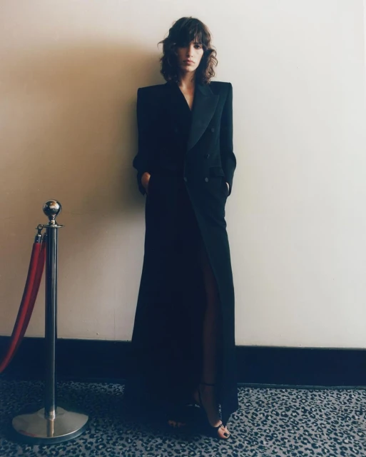 A woman in a long, dark coat poses against a neutral wall next to a silver bollard with a red rope, showcasing a fashion-editorial style.