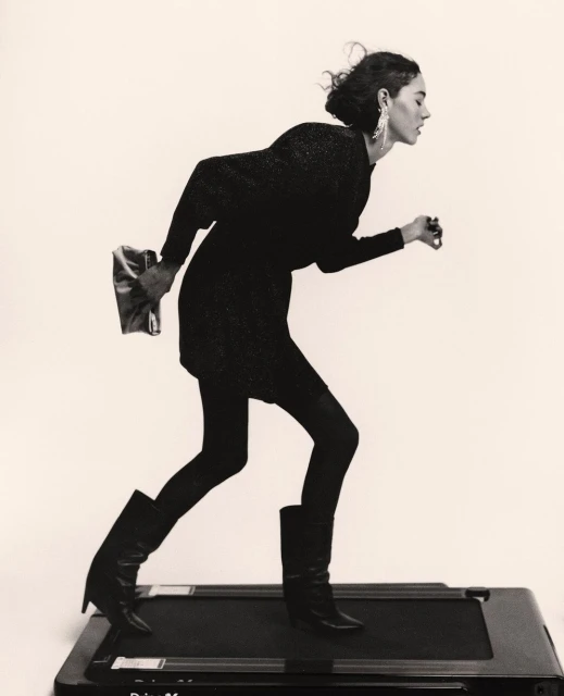 Monochrome portrait of a woman in a dress on a treadmill, holding a clutch, side profile view, in a photo studio against a plain backdrop.