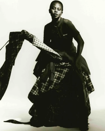 Elegant, dark-skinned woman posing in a dramatic layered plaid dress against a bright background, fashion-forward and artistic.