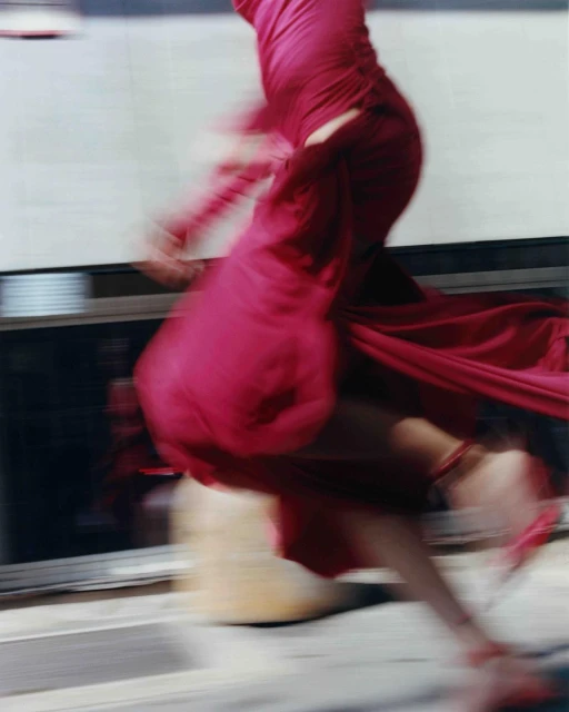 Fashion photography featuring a blurry, moving figure in a bright pink dress and matching high heels on a sidewalk, conveying motion and style.