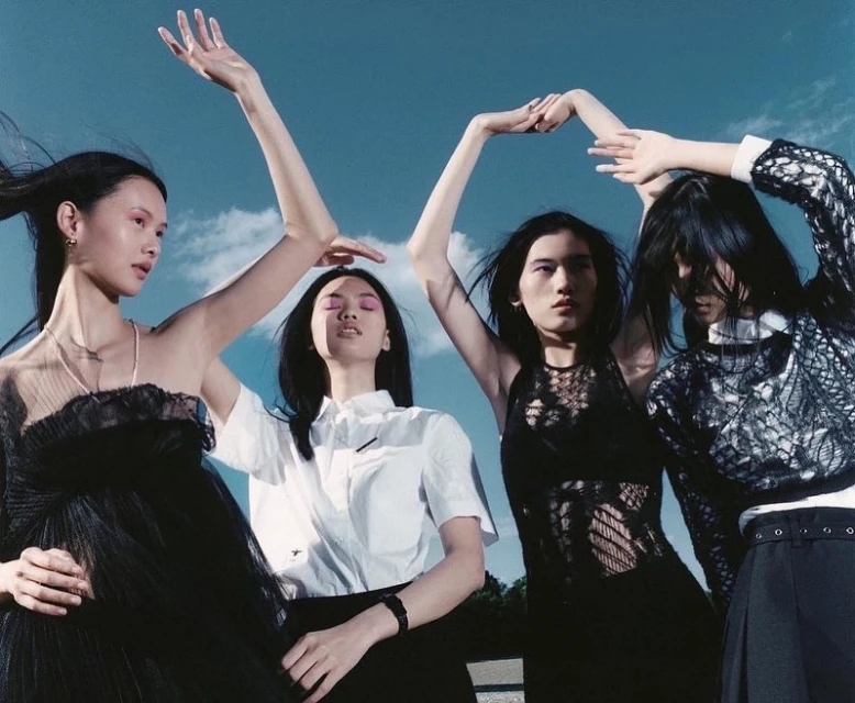 Four Asian women in black and white fashions are posed outdoors against a blue sky, two with arms raised in a coordinated high fashion shot.