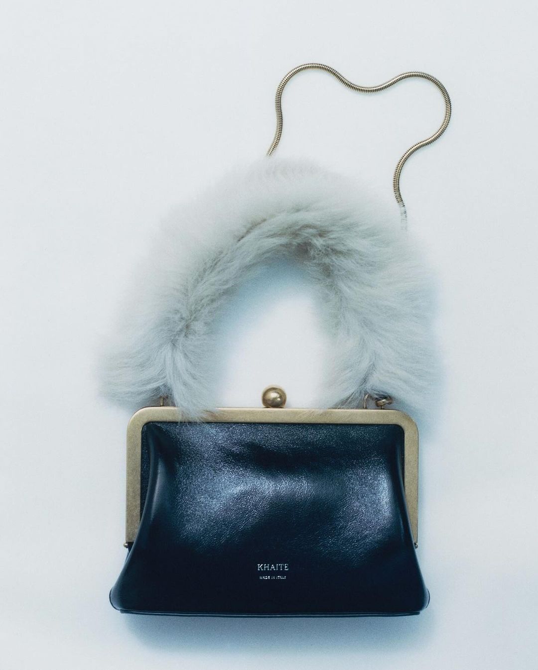A black leather handbag with a plush white faux-fur handle and a thin golden metal chain strap against a plain white background.