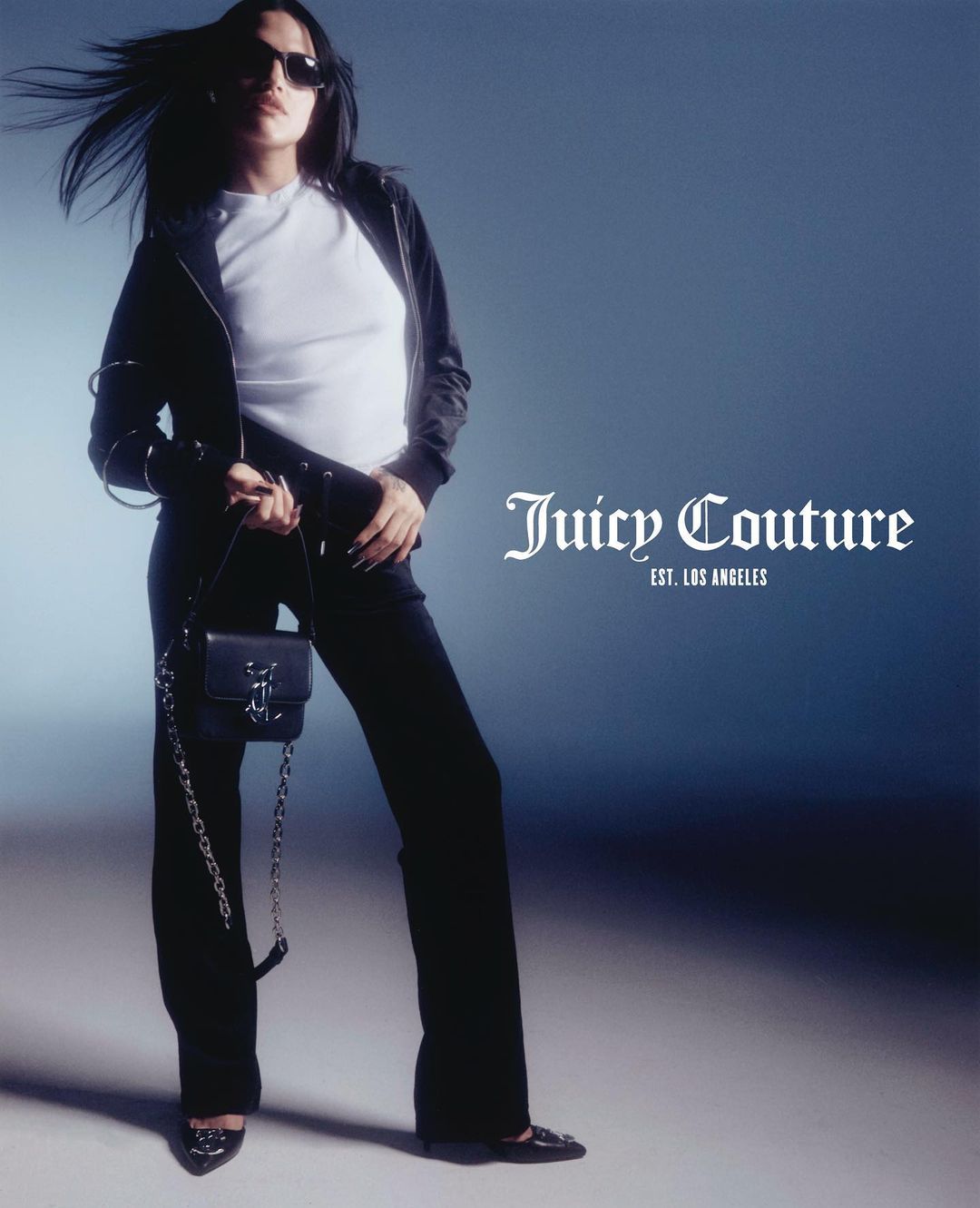 A brunette adult woman in black wide-leg pants, jacket, and a white t-shirt, holding a black chain-strap purse in a studio setting.