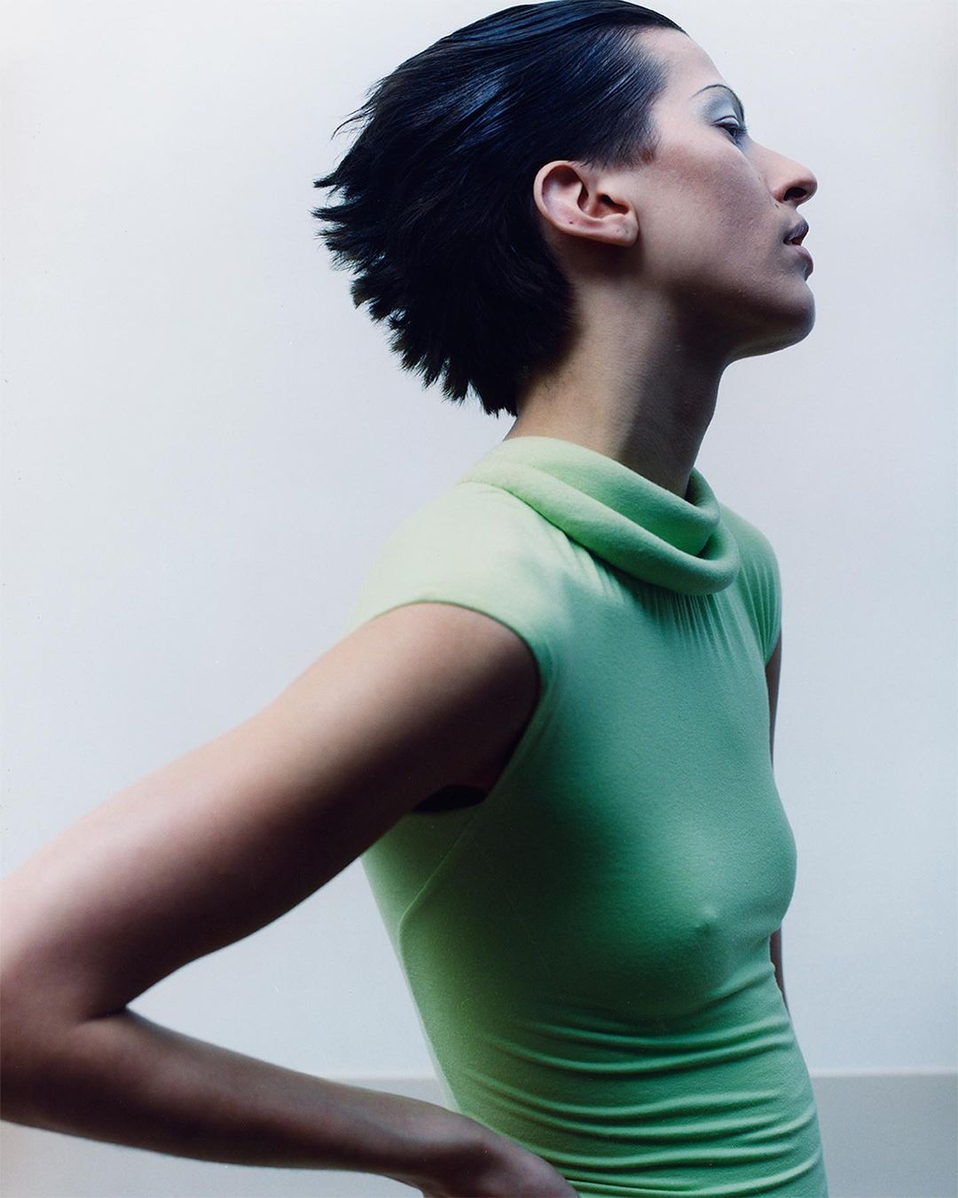 Profile of a woman with short black hair, wearing a light green turtleneck dress against a plain white backdrop, posing with hands on hips.