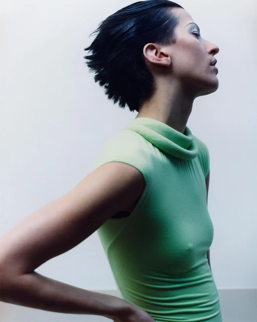 Profile of a woman with short black hair, wearing a light green turtleneck dress against a plain white backdrop, posing with hands on hips.