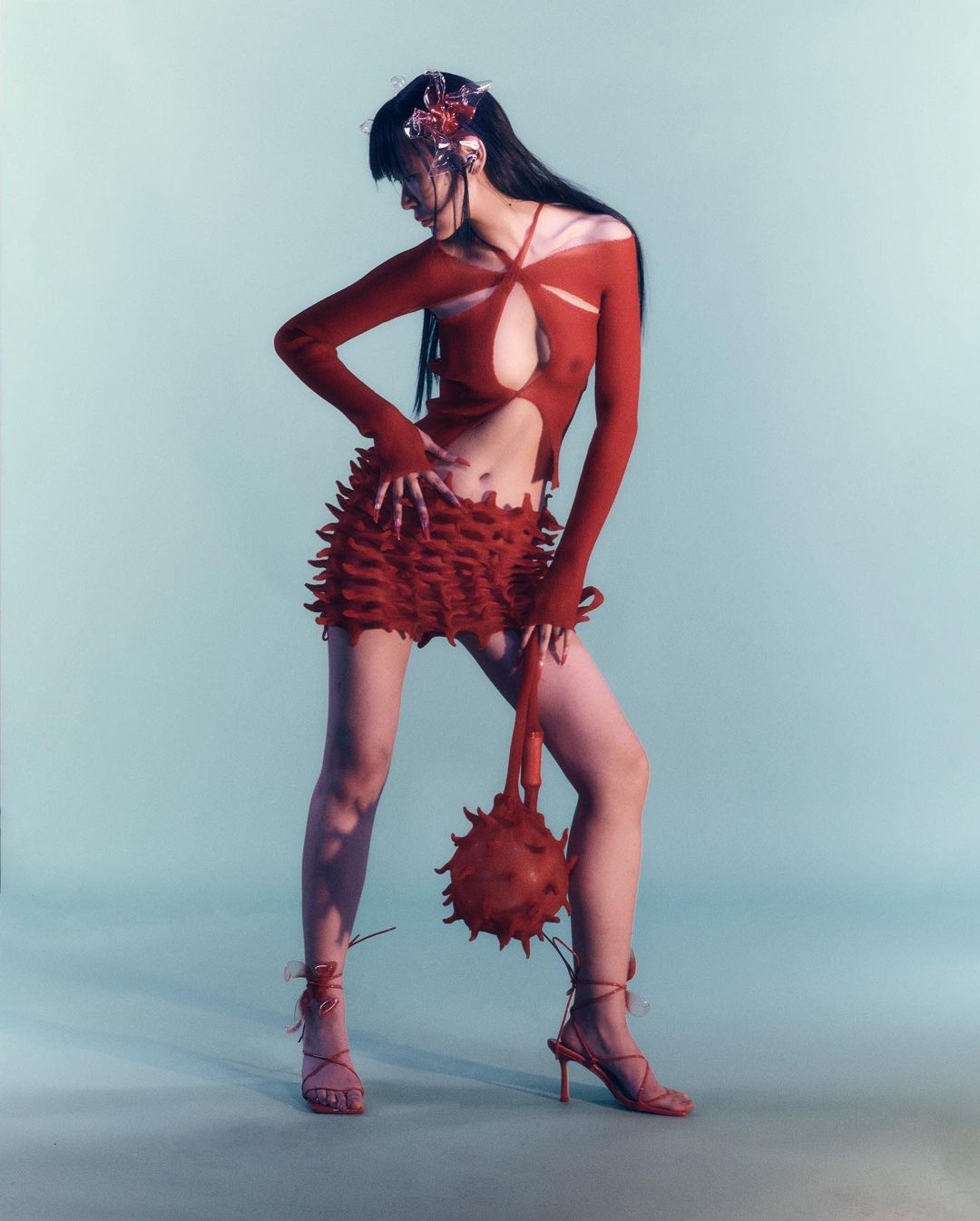 A model poses in a red cut-out top and ruffled skirt with spiked sphere purse against a light blue backdrop in a studio shot.