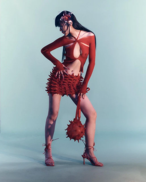 A model poses in a red cut-out top and ruffled skirt with spiked sphere purse against a light blue backdrop in a studio shot.