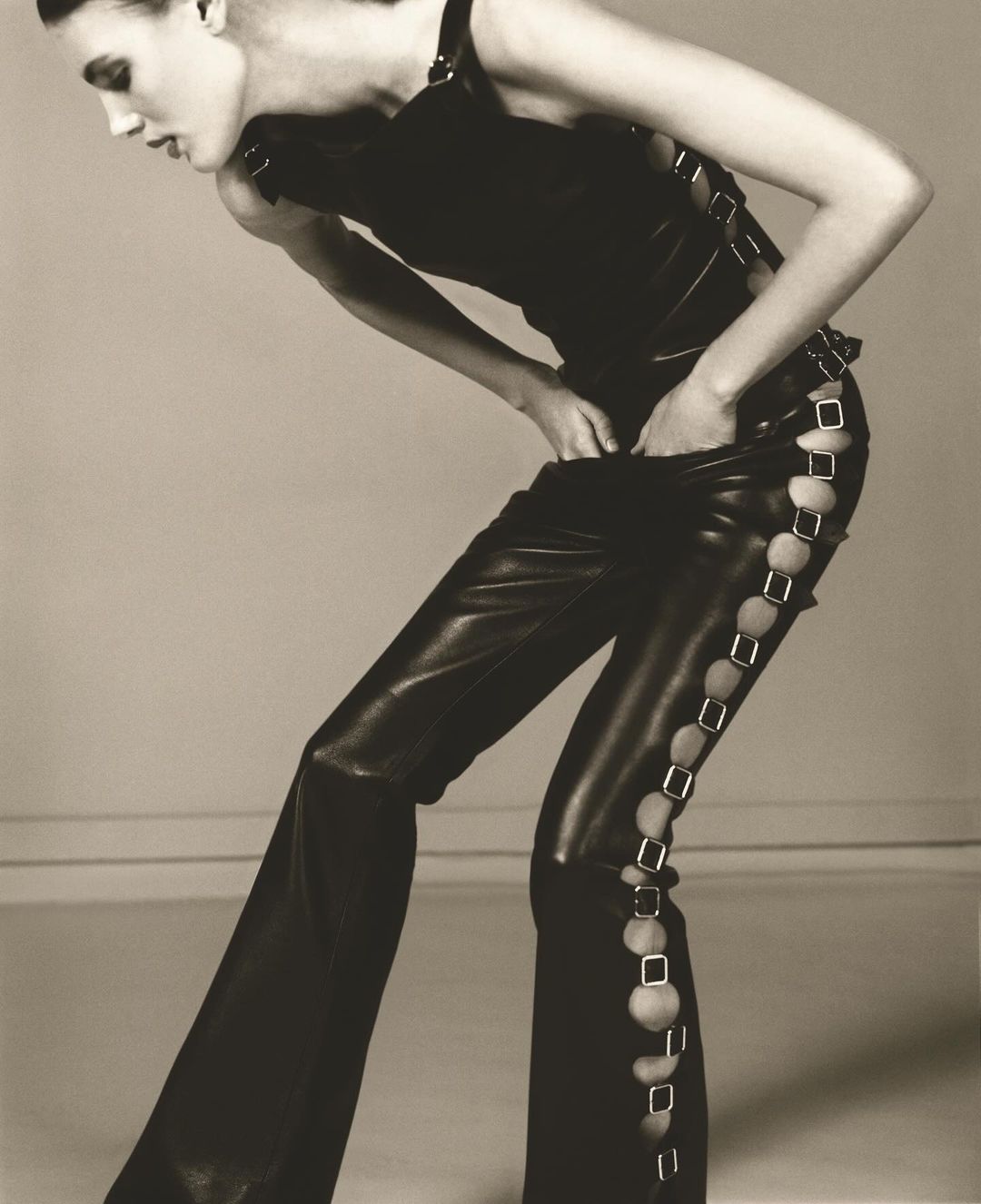 Fashion model poses in a black top and leather pants with decorative cutouts against a neutral background in a studio photograph.
