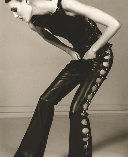 Fashion model poses in a black top and leather pants with decorative cutouts against a neutral background in a studio photograph.
