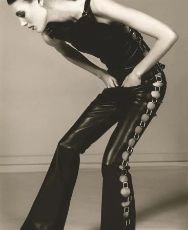 Fashion model poses in a black top and leather pants with decorative cutouts against a neutral background in a studio photograph.