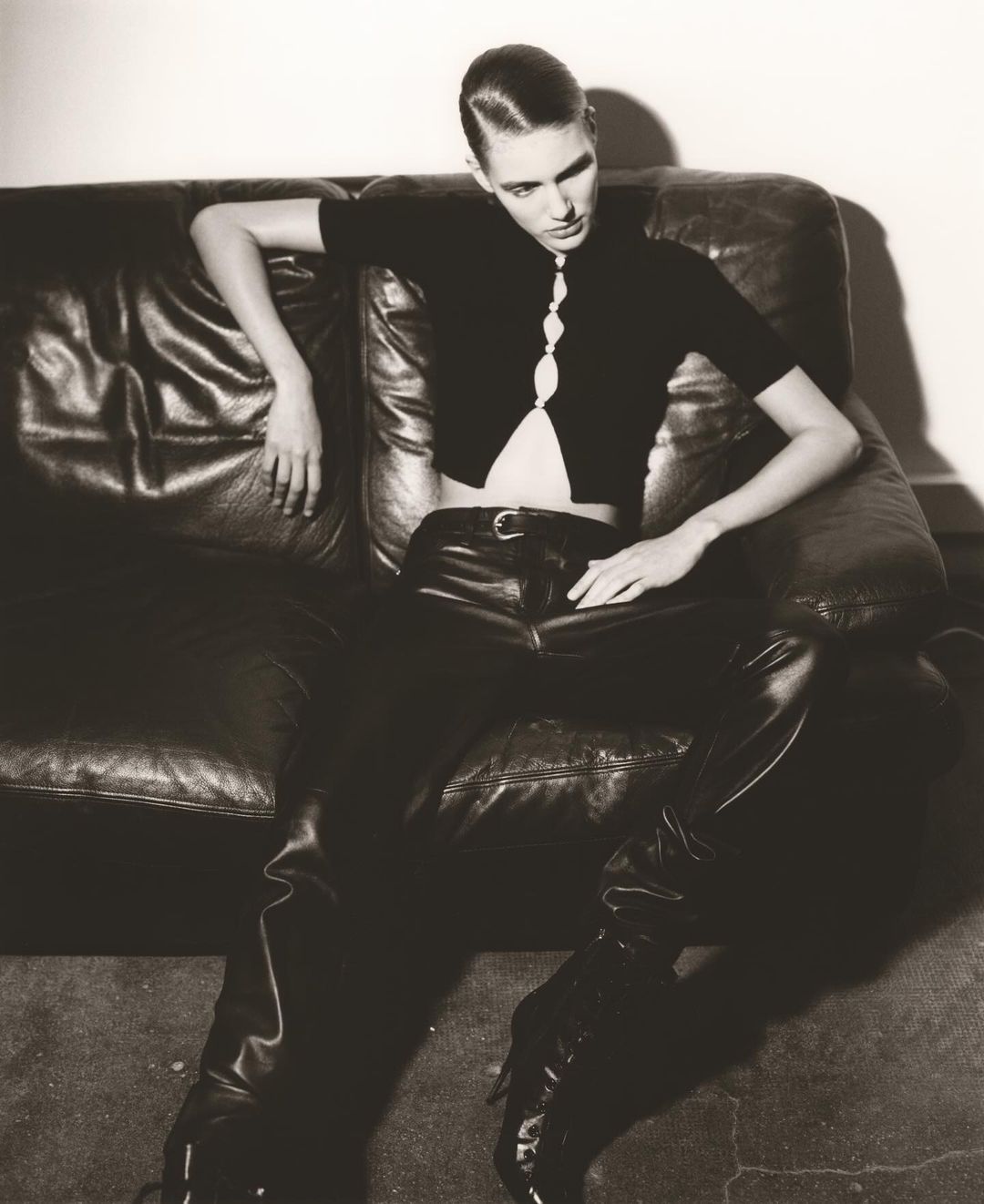 A young adult woman in black leather pants and a knit top poses on a dark leather couch in an indoor setting with a neutral background.