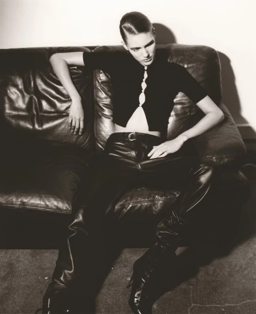 A young adult woman in black leather pants and a knit top poses on a dark leather couch in an indoor setting with a neutral background.