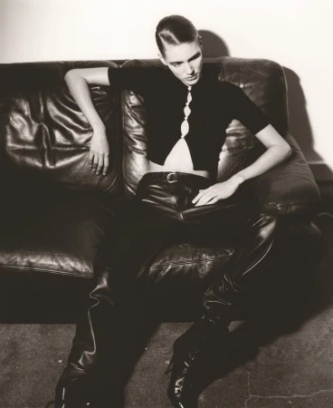 A young adult woman in black leather pants and a knit top poses on a dark leather couch in an indoor setting with a neutral background.