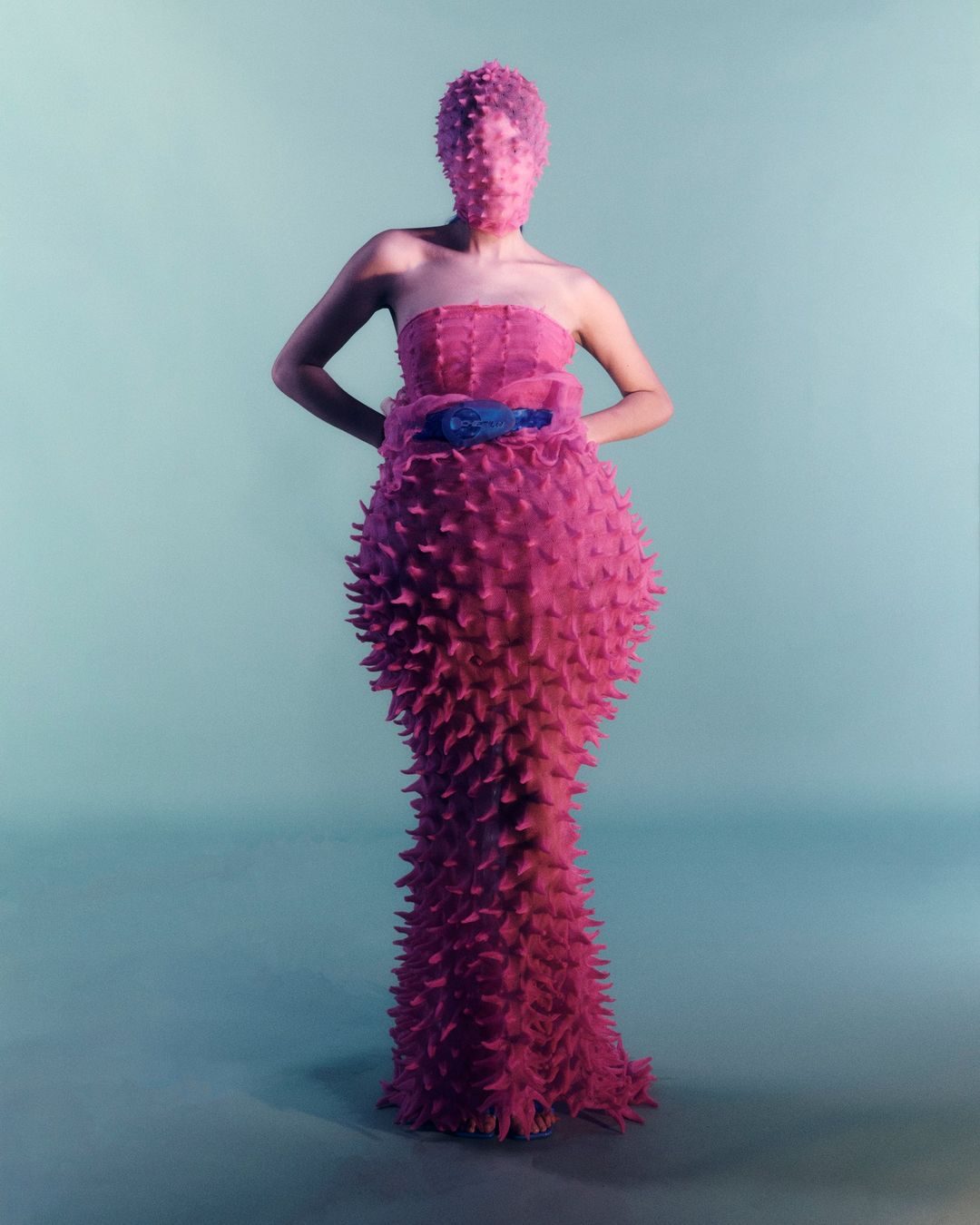 A person wearing a pink spiky gown and head covering stands in a studio against a blue background for a fashion editorial photoshoot.