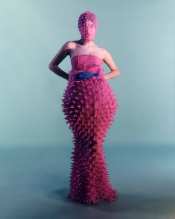 A person wearing a pink spiky gown and head covering stands in a studio against a blue background for a fashion editorial photoshoot.