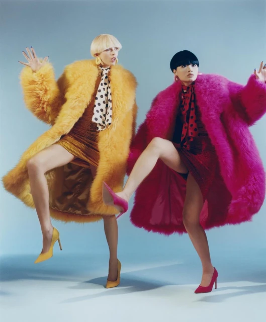 Two fashion models in vibrant faux fur coats and heels strike playful poses against a light blue backdrop in a studio setting.