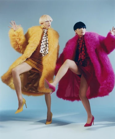 Two fashion models in vibrant faux fur coats and heels strike playful poses against a light blue backdrop in a studio setting.