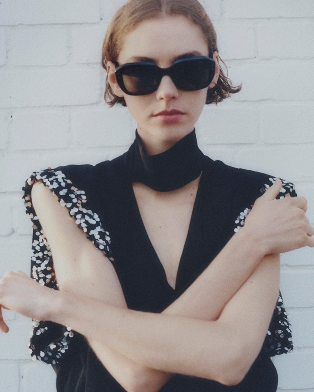 Model wearing dark sunglasses and a black top with sequined shoulders against a white brick wall