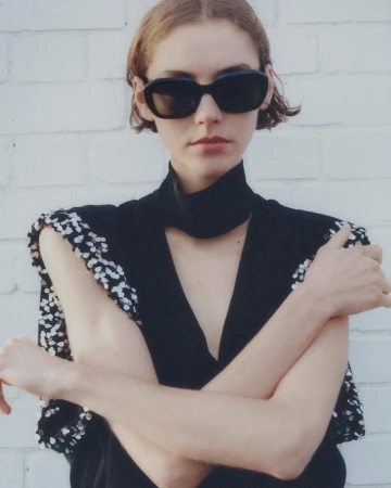 Model wearing dark sunglasses and a black top with sequined shoulders against a white brick wall