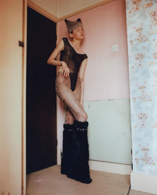 A slender person with light skin stands in an outfit with lace tights and fuzzy boots in a room with teddy bear wallpaper.