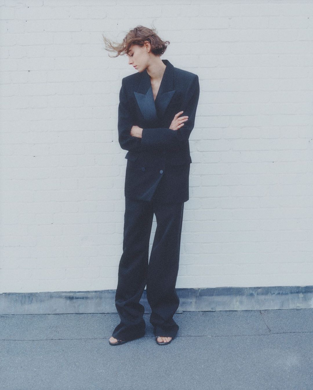 A solitary young woman in a black suit stands with crossed arms against a white brick wall, showcasing editorial fashion.