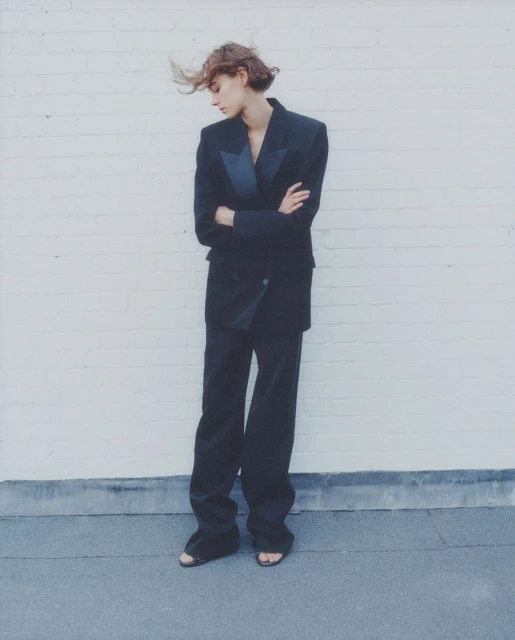 A solitary young woman in a black suit stands with crossed arms against a white brick wall, showcasing editorial fashion.