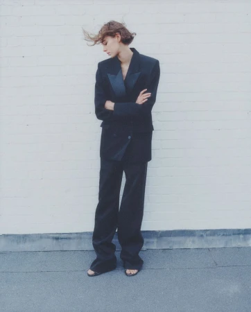 A solitary young woman in a black suit stands with crossed arms against a white brick wall, showcasing editorial fashion.