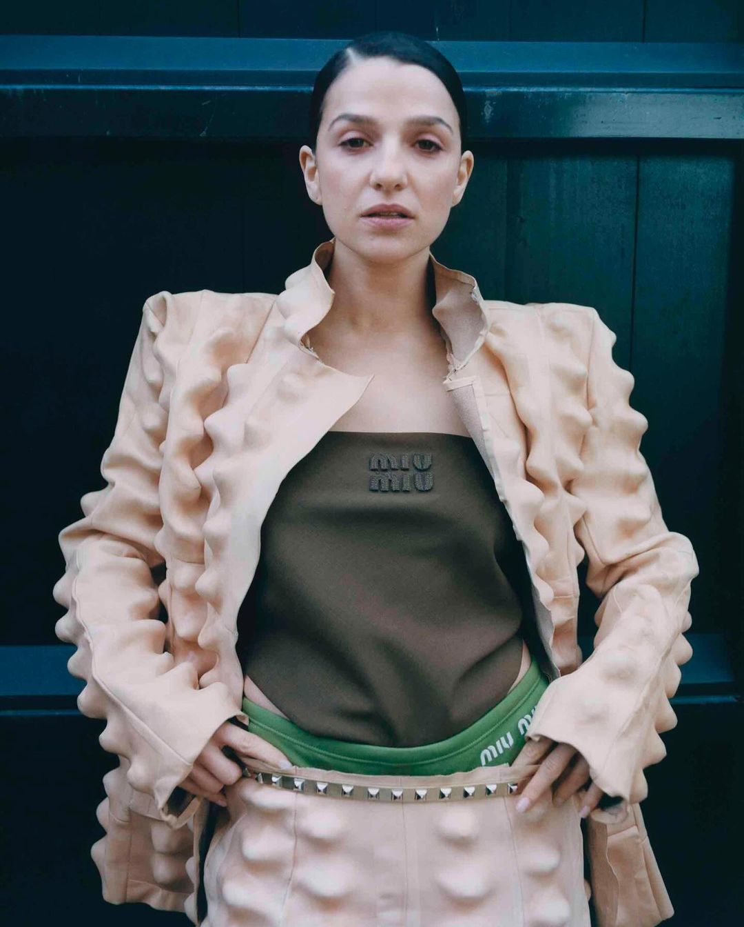 Woman in avant-garde fashion wearing Miu Miu crop top and skirt, standing against a dark wall in a medium studio shot, neutral expression.