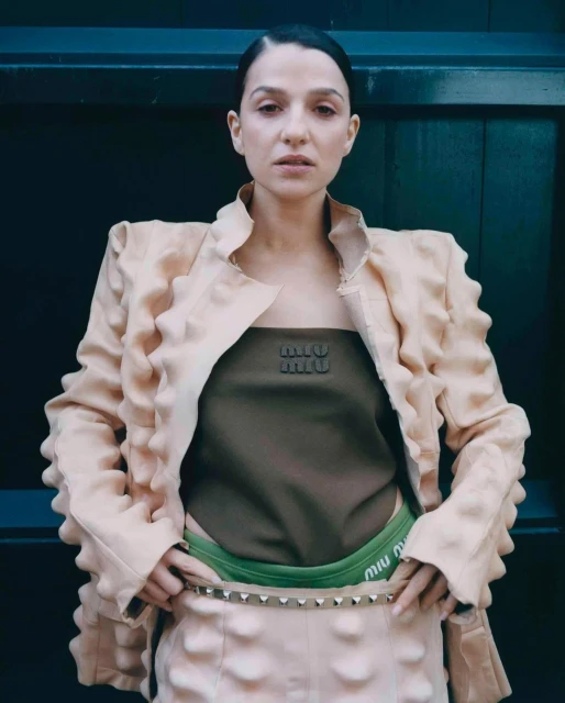 Woman in avant-garde fashion wearing Miu Miu crop top and skirt, standing against a dark wall in a medium studio shot, neutral expression.