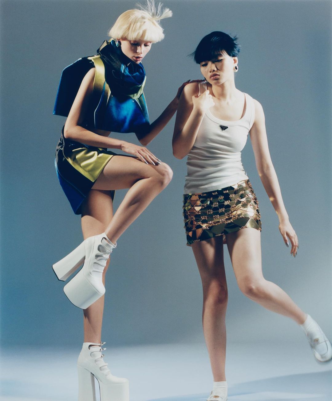 Two women in fashionable outfits and platform heels pose in a studio, showcasing contemporary design against a gradient blue background.