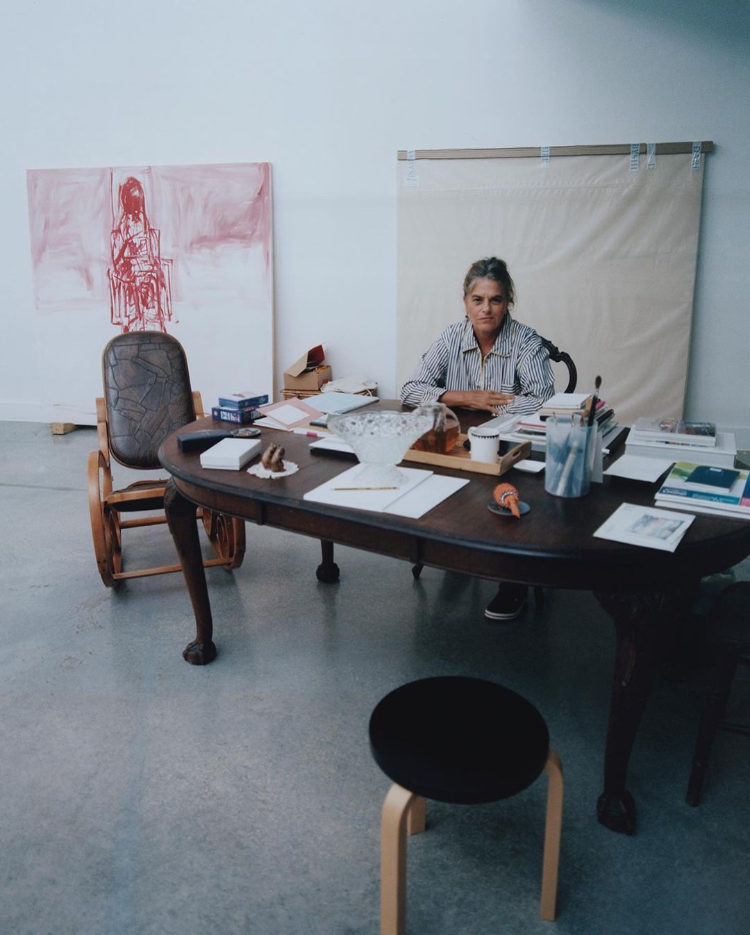 Woman seated at a table laden with papers and objects, working in a bright studio with paintings and stool nearby.