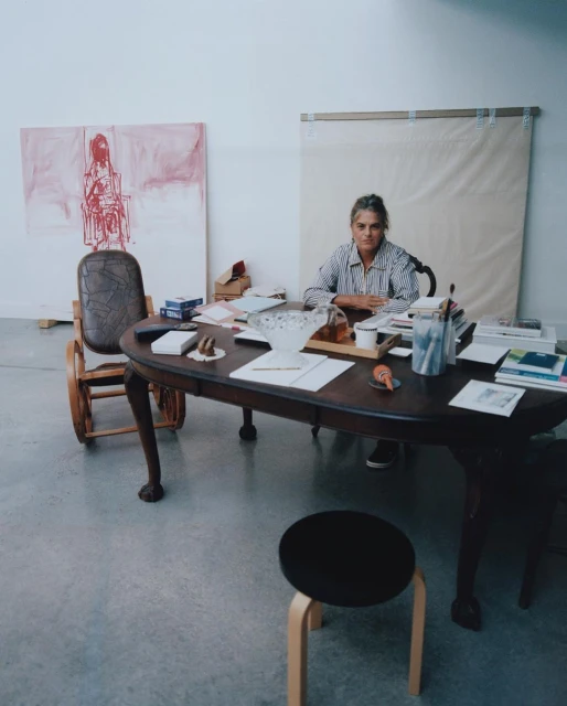 Woman seated at a table laden with papers and objects, working in a bright studio with paintings and stool nearby.