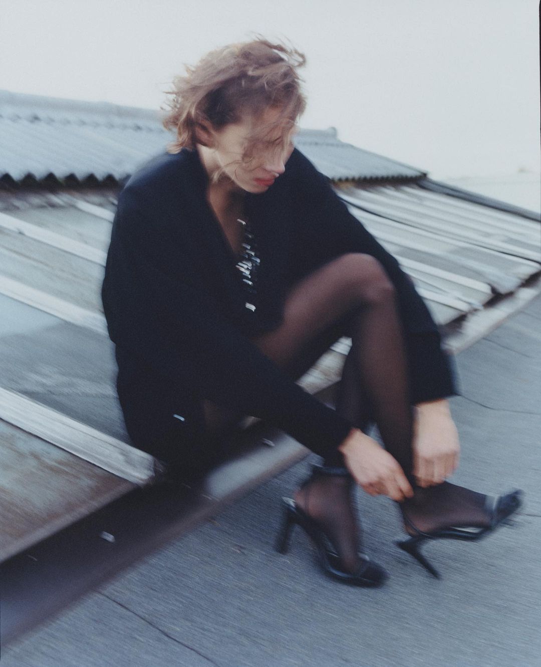 Woman in black hosiery and heels sits on a rooftop adjusting her shoe in overcast weather. She is wearing a blazer and has short brown hair.