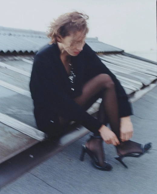 Woman in black hosiery and heels sits on a rooftop adjusting her shoe in overcast weather. She is wearing a blazer and has short brown hair.