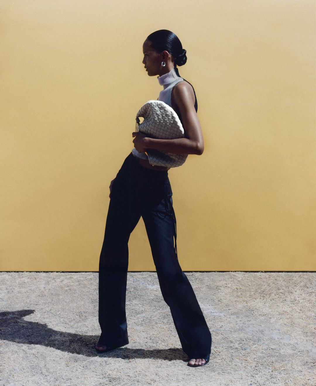 A woman in black pants and a gray top poses with a woven bag against a sunny yellow backdrop, bathed in bright, soft lighting.