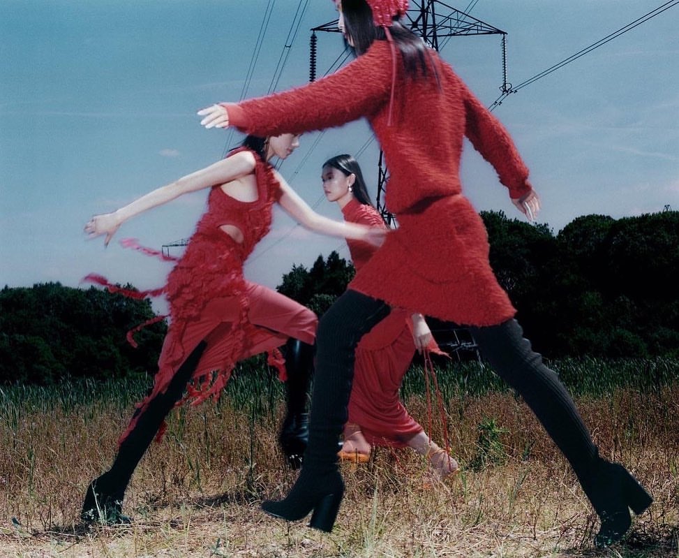 Three fashion models in red outfits and black boots pose in a grassy field under a blue sky with electrical towers, creating a surreal outdoor fashion shot.
