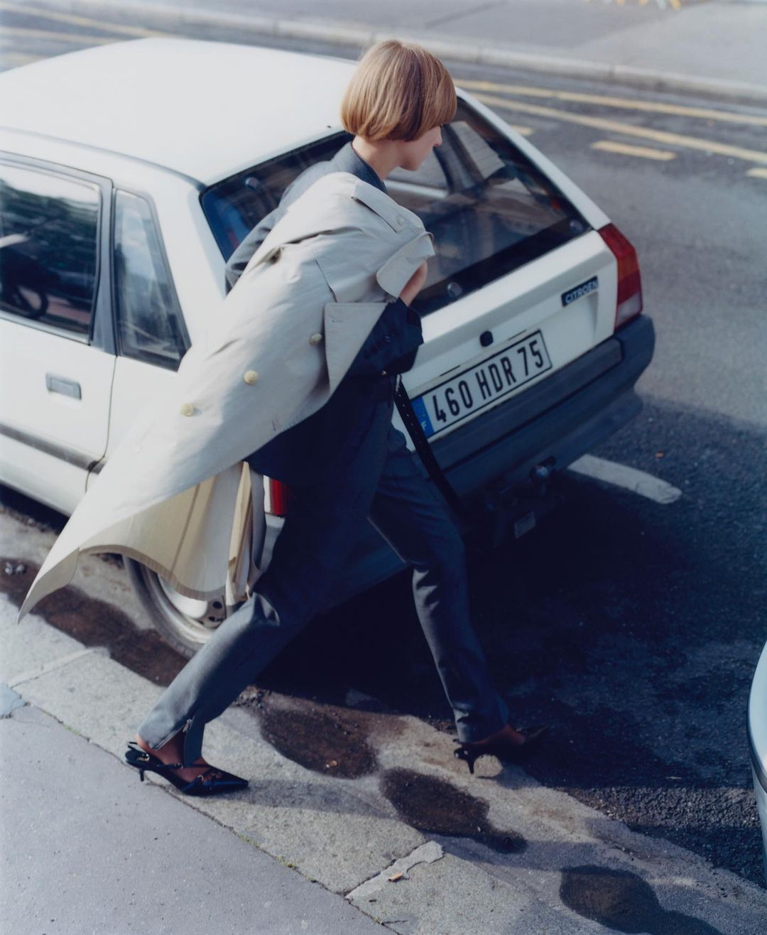 Model with bob haircut walking past a white vintage hatchback car on a city street, wearing a cream coat over a dark suit.