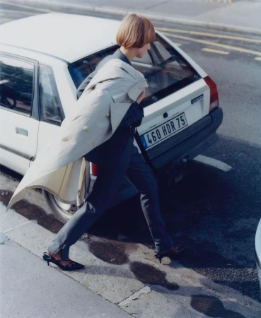 Model with bob haircut walking past a white vintage hatchback car on a city street, wearing a cream coat over a dark suit.