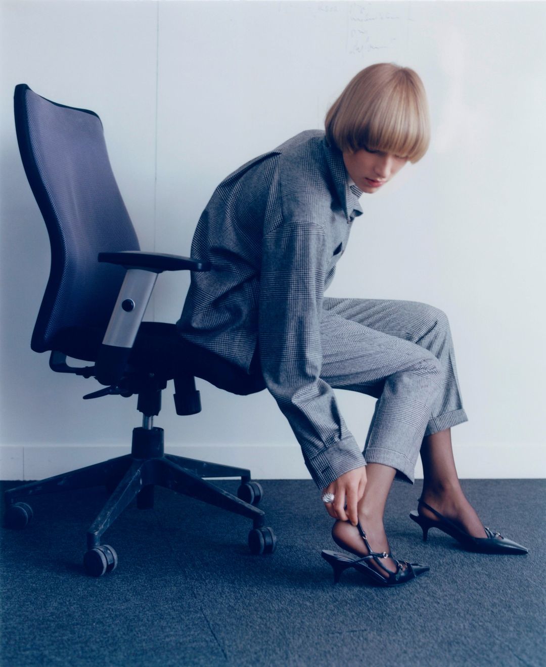 Woman in grey suit adjusts black heeled sandal while seated in office chair against a white wall and blue carpet.