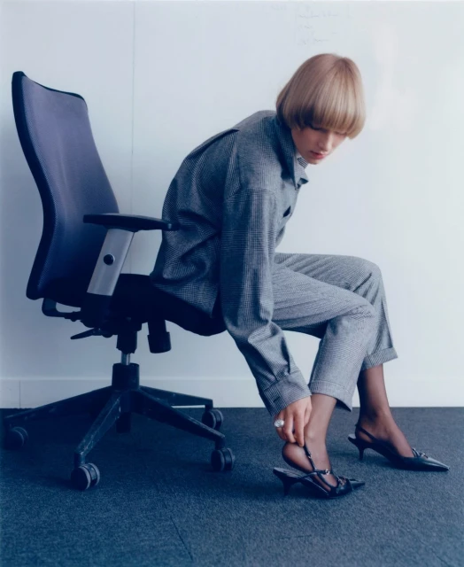 Woman in grey suit adjusts black heeled sandal while seated in office chair against a white wall and blue carpet.
