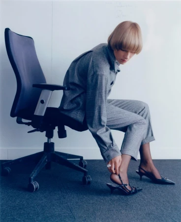 Woman in grey suit adjusts black heeled sandal while seated in office chair against a white wall and blue carpet.