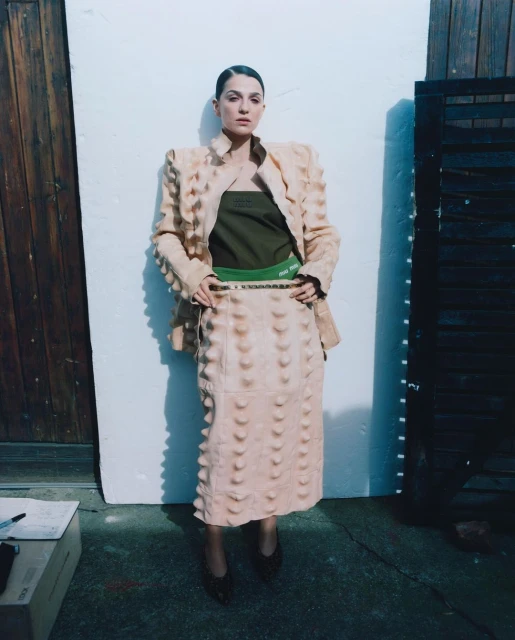 A woman in a light peach jacket and skirt with decorative bumps poses against a wall with a wooden door and gate in the background.