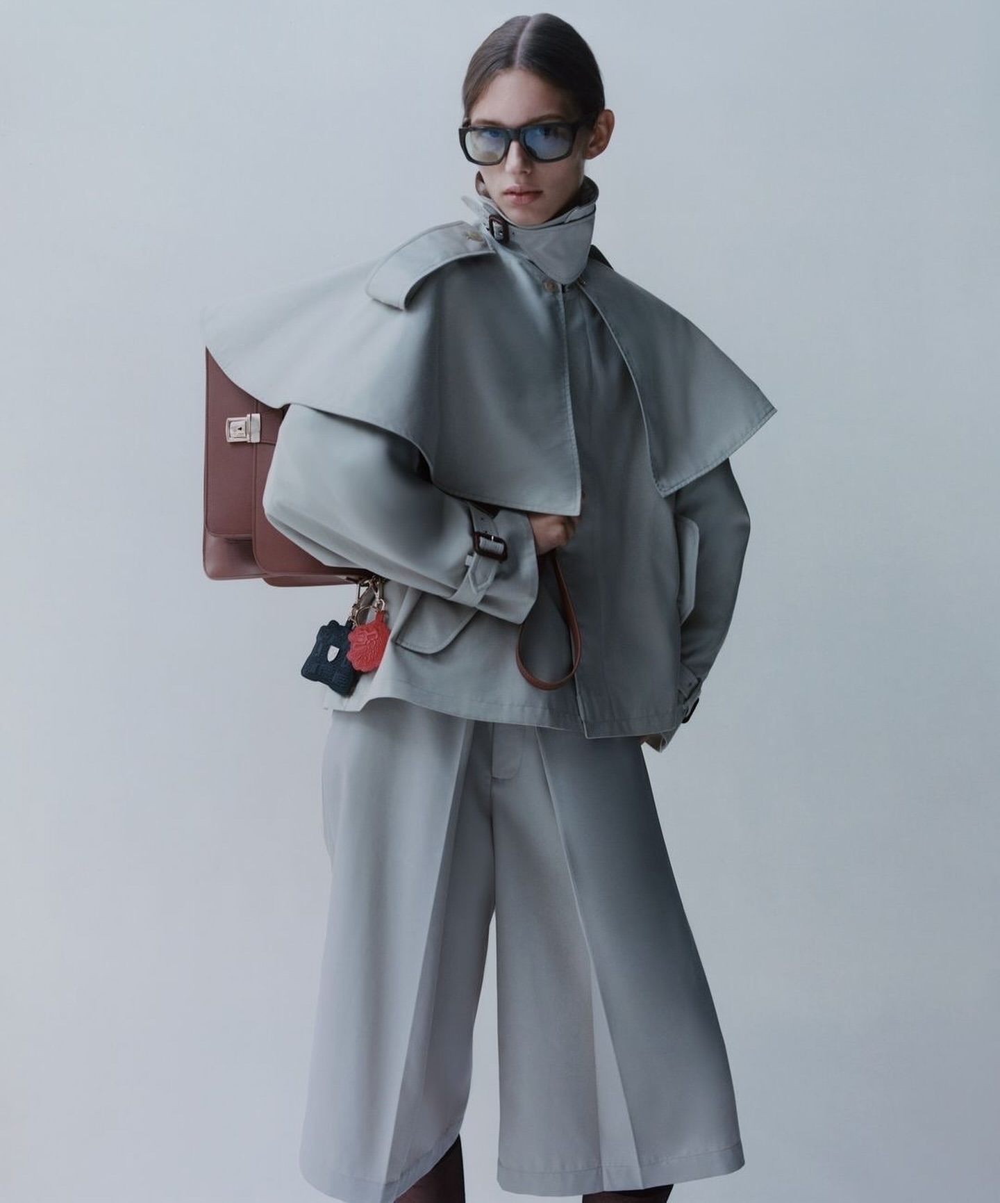 Fashion editorial featuring a female model in a grey trench coat and culottes, posing with a brown leather briefcase and dark sunglasses.