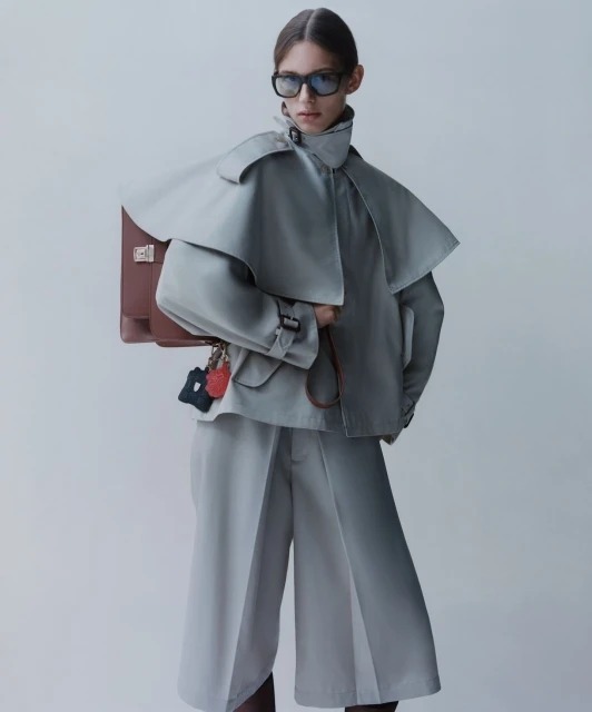 Fashion editorial featuring a female model in a grey trench coat and culottes, posing with a brown leather briefcase and dark sunglasses.