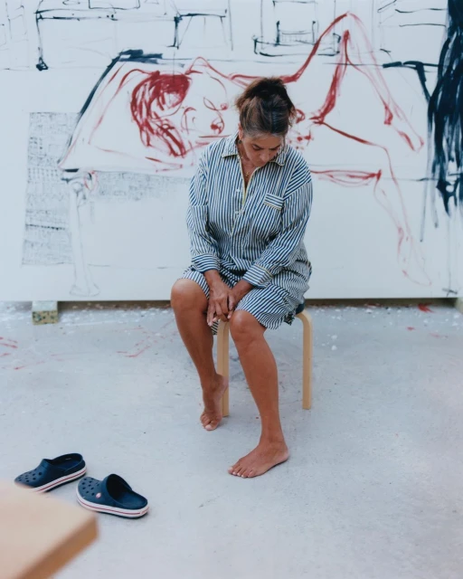 Artist in striped shirt sits on a stool in her studio in front of a large canvas with a red drawing; a pair of clogs on floor.