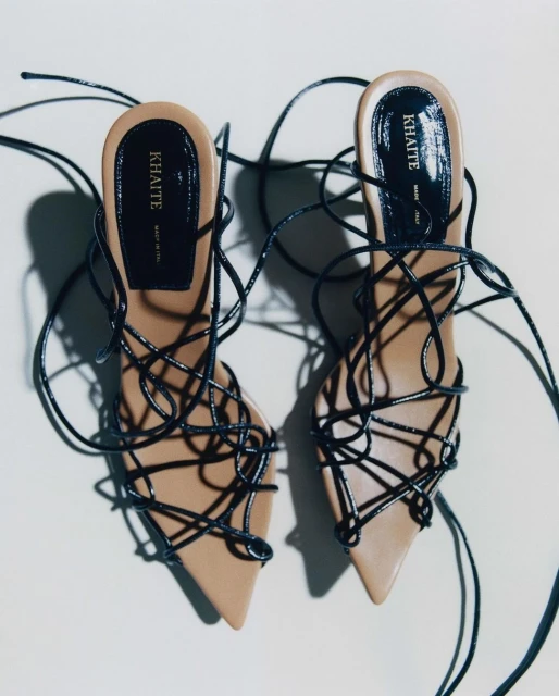 Elegant pair of beige leather heeled sandals with black wraparound straps, displayed on a seamless white background. Fashion-forward footwear.