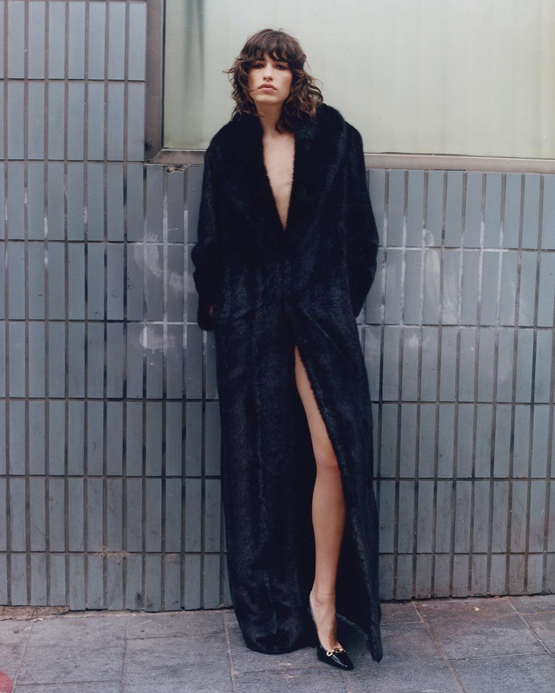 Adult woman posing in a long black fur coat with a thigh-high slit, wearing black heels against a gray tiled wall outdoors.